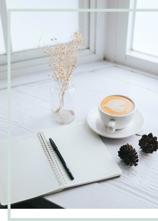 a desk with a notebook, pen, coffee, and a vase with dried flowers.